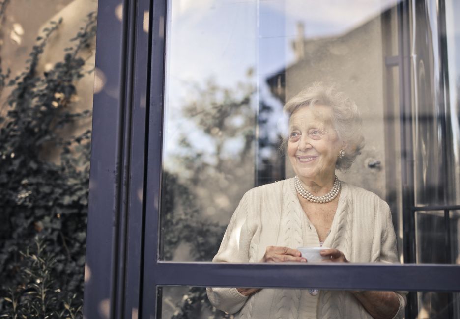Elderly woman smiles by a glass window, holding a cup, in a peaceful setting.