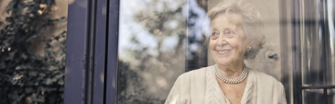 Elderly woman smiles by a glass window, holding a cup, in a peaceful setting.